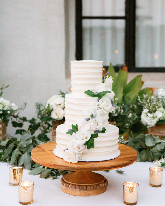 White wedding cake with floral decorations on a wooden stand, surrounded by greenery and candles.